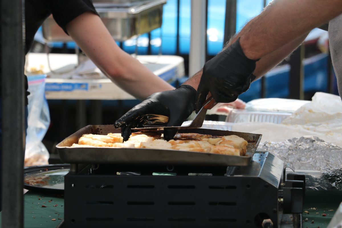 A man makes grilled cheese on a small burner while a person behind him prepared a carrier for the food.