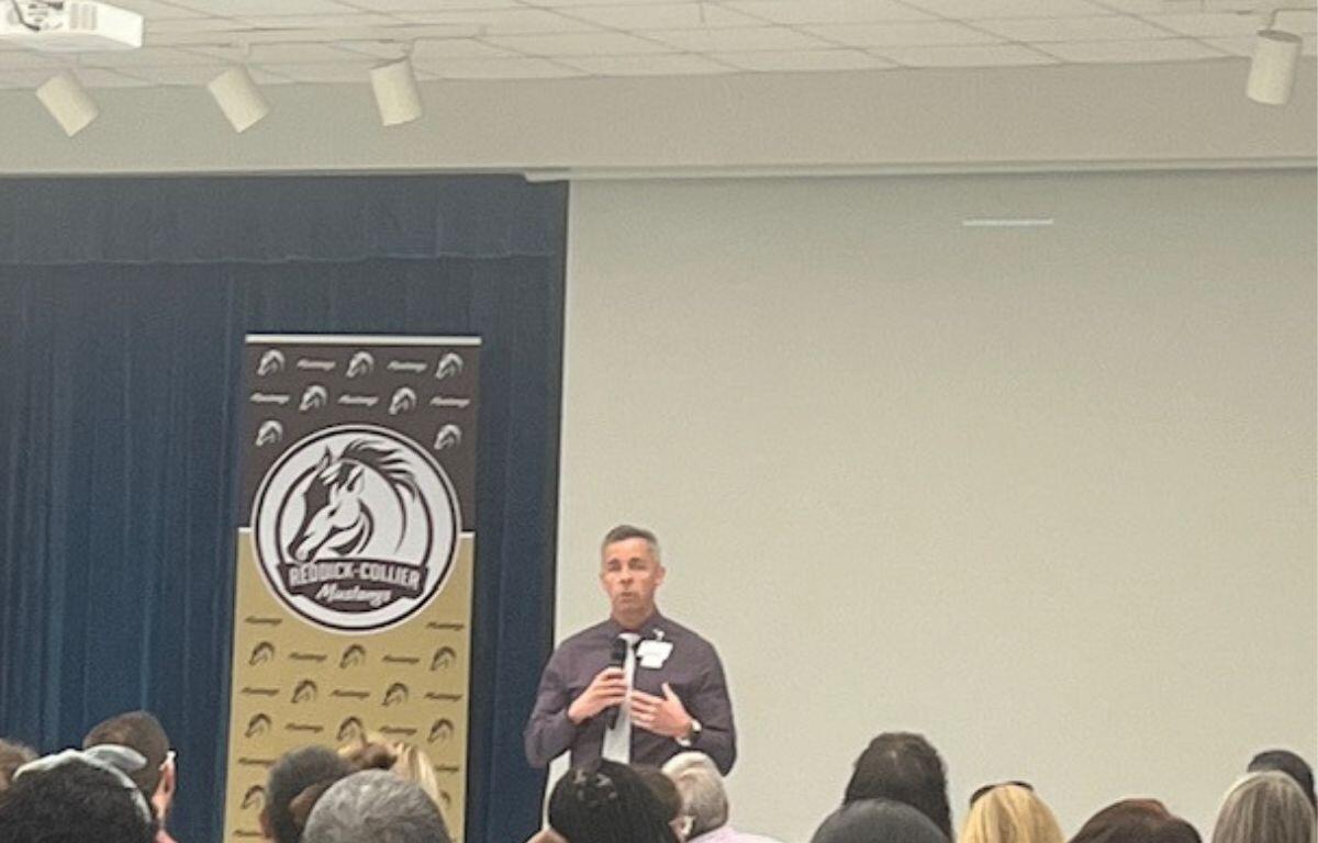 Blue curtain to the left in the background, multi-colored placard with an insignia and writing and to the right an off-white colored wall. A man speaking and the back of people's heads in the foregrounds.