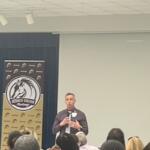 Blue curtain to the left in the background, multi-colored placard with an insignia and writing and to the right an off-white colored wall. A man speaking and the back of people's heads in the foregrounds.