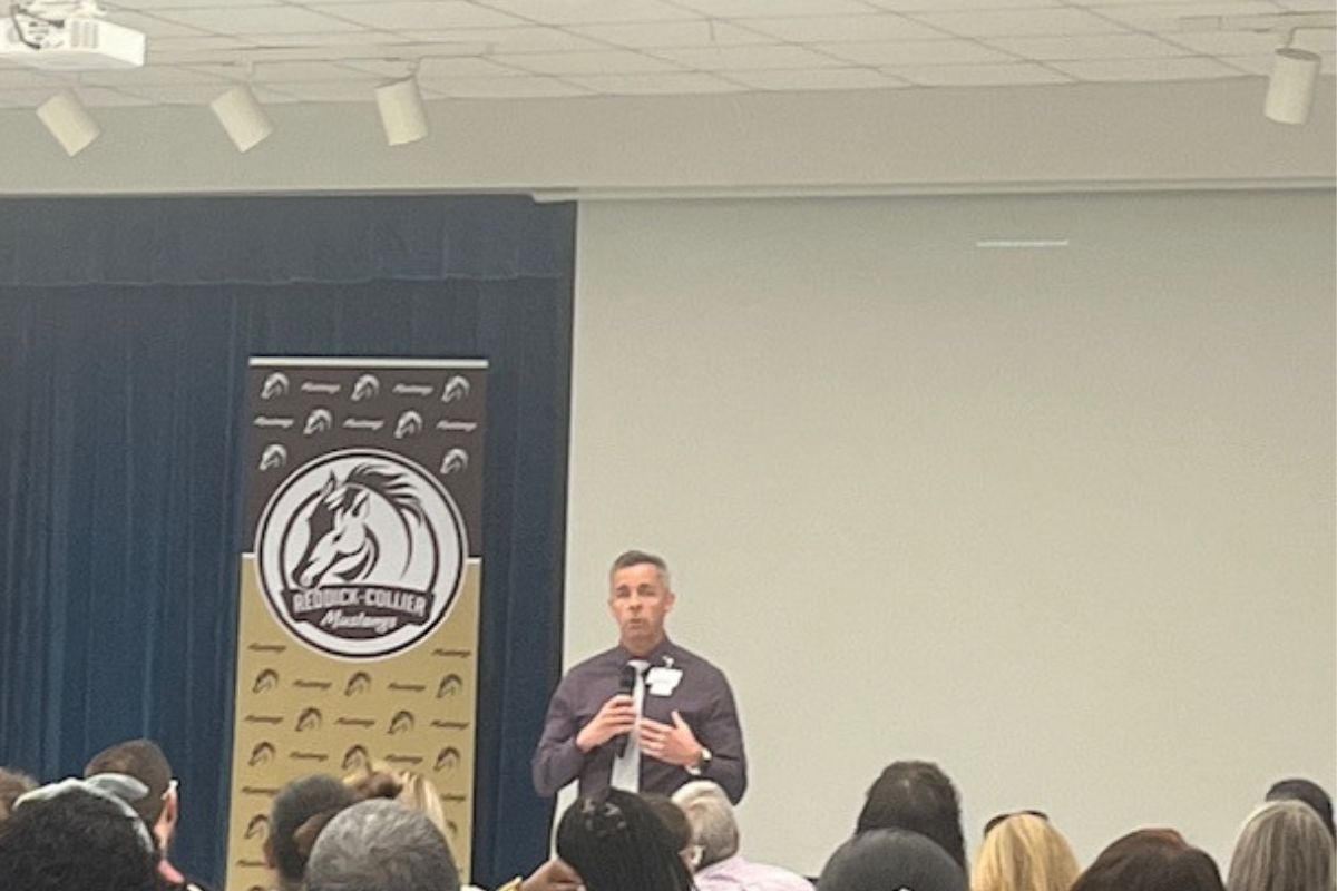 Blue curtain to the left in the background, multi-colored placard with an insignia and writing and to the right an off-white colored wall. A man speaking and the back of people's heads in the foregrounds.