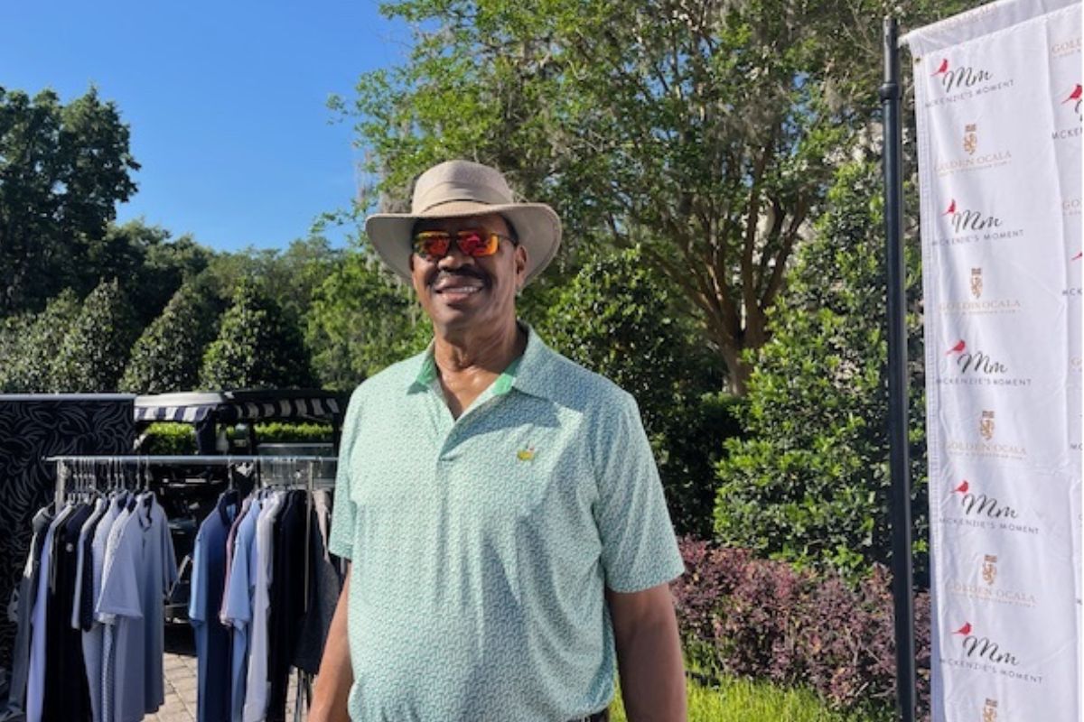 A man wearing a hat and sunglass, with a rack of shirts to his left, a fixture holding a backdrop to his right, trees and a tent in the background. 