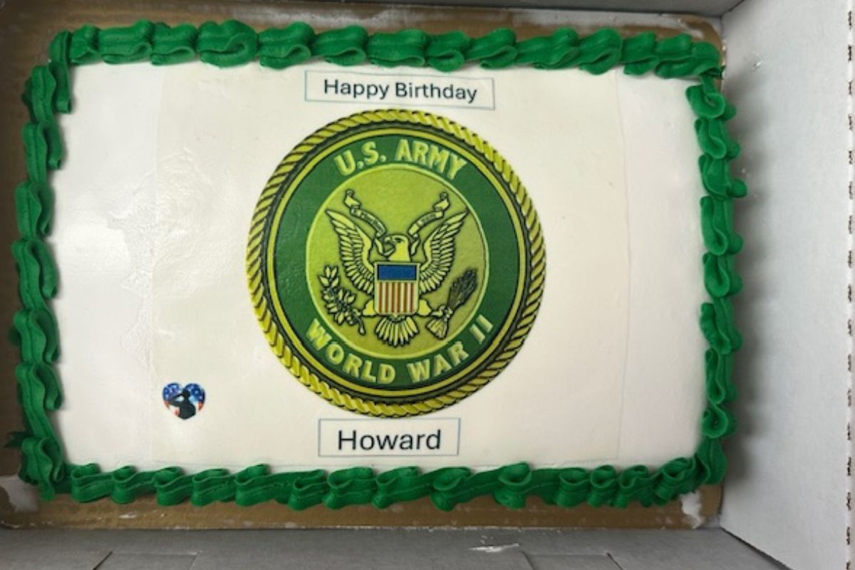 A birthday cake, with green trim, and an insignia with writing in the center, with white background. 