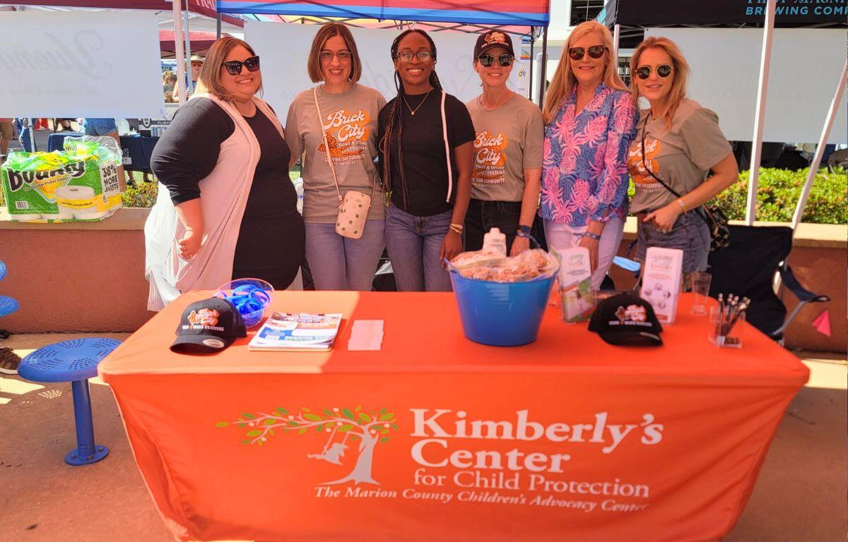 A group of women stand behind a table with a bright orange tablecloth with words reading, "Kimberly's Center for Child Protection."