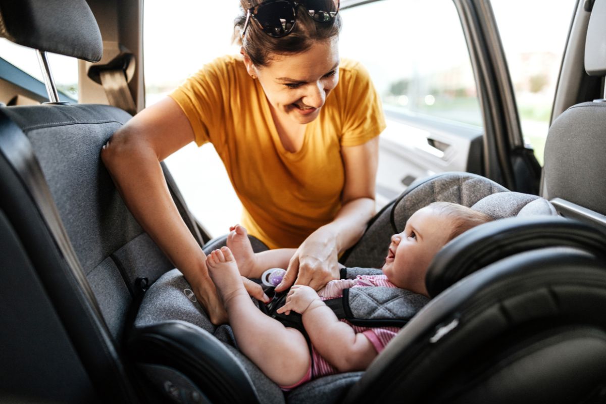 Colored image of a women in an orange shirt strapping a children into a car seat.