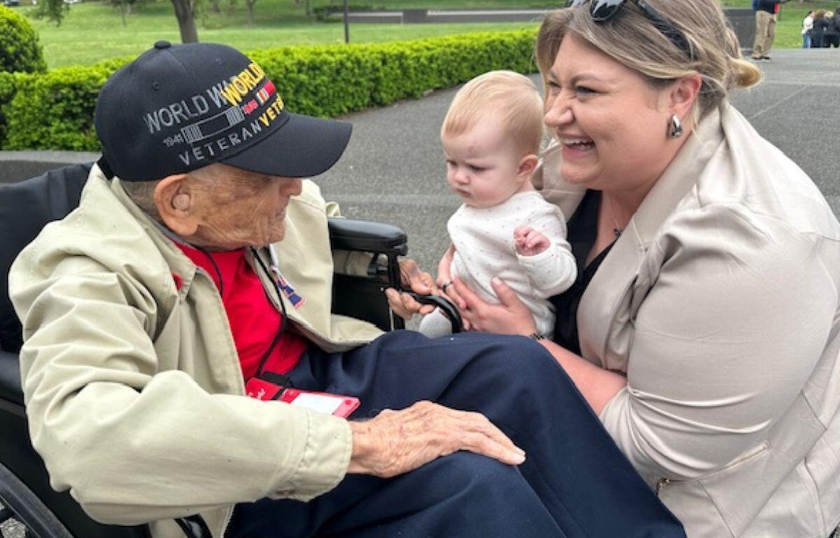 A man in a wheelchair and baseball hat talks with a woman, holding an infant, with a walkway and hedges in the background.