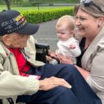 A man in a wheelchair and baseball hat talks with a woman, holding an infant, with a walkway and hedges in the background.