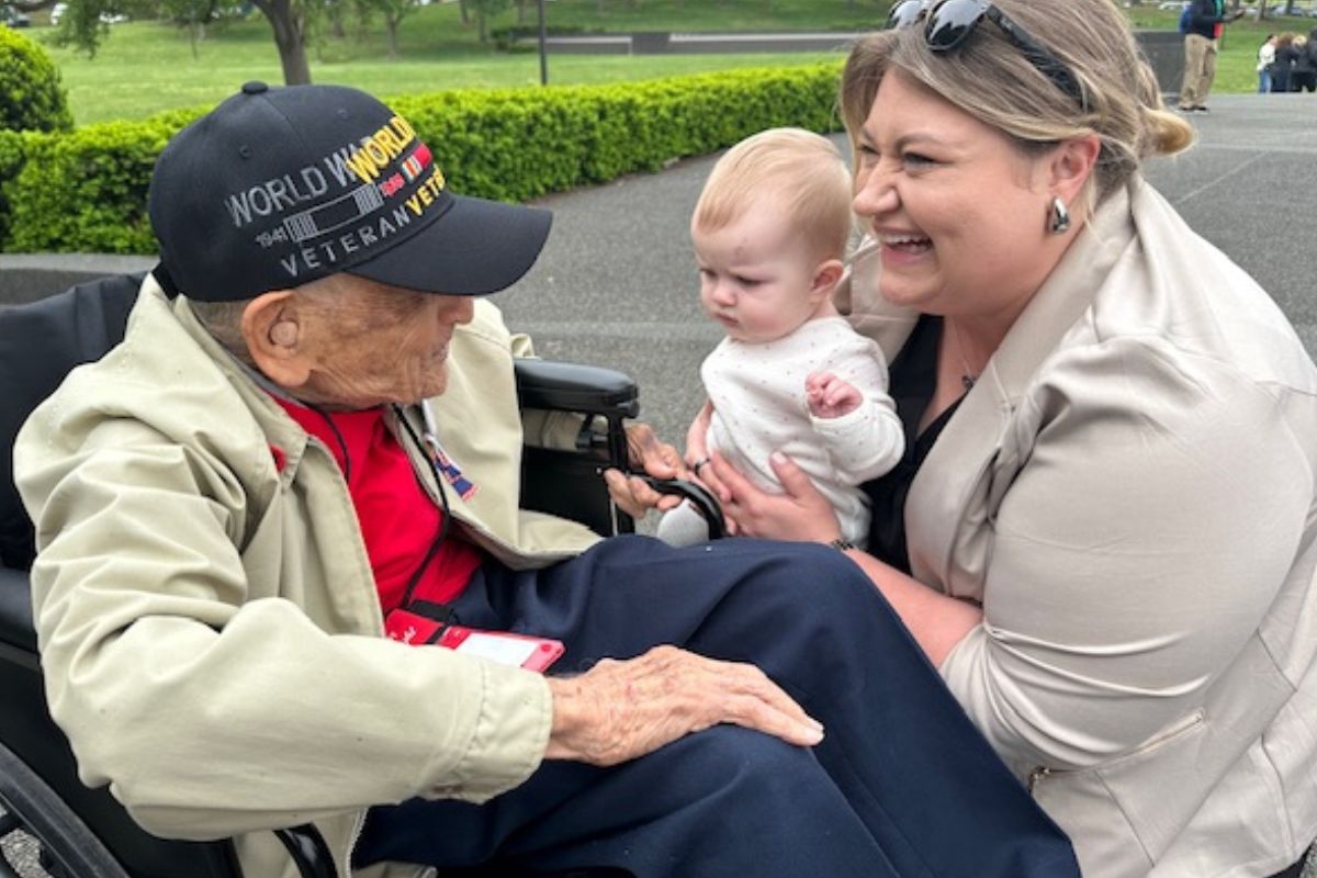 A man in a wheelchair and baseball hat talks with a woman, holding an infant, with a walkway and hedges in the background.