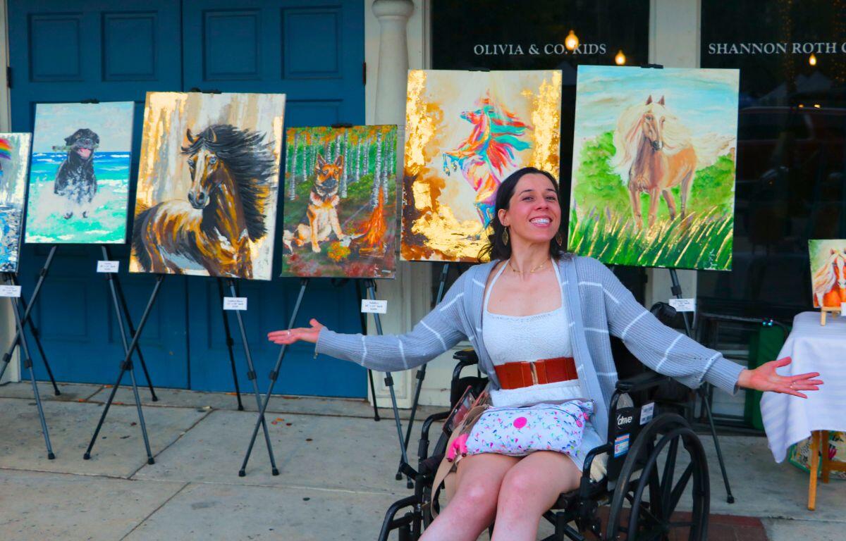 A woman sits in a wheelchair in front of a line of paintings, gesturing to them.