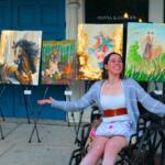 A woman sits in a wheelchair in front of a line of paintings, gesturing to them.
