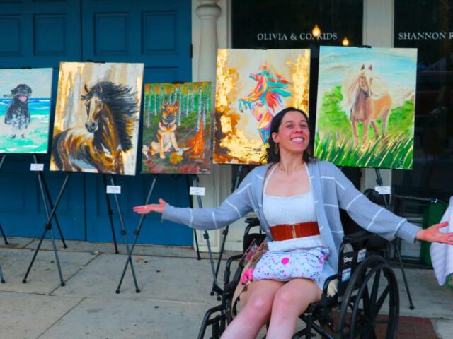 A woman sits in a wheelchair in front of a line of paintings, gesturing to them.