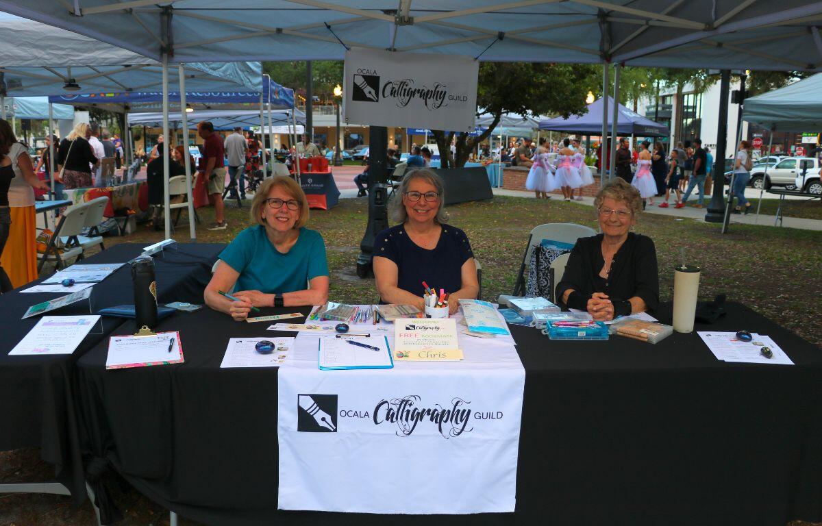 Three women sit behind a black table with a white table runner reading, "Ocala Calligraphy Guild." The table featured sign-up sheets and calligraphy activities.
