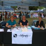 Three women sit behind a black table with a white table runner reading, "Ocala Calligraphy Guild." The table featured sign-up sheets and calligraphy activities.