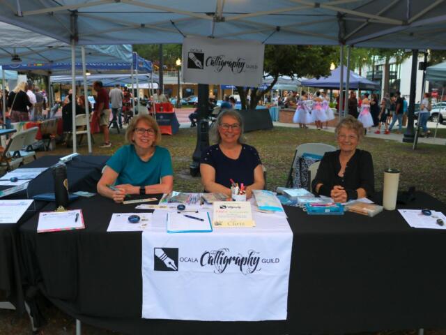 Three women sit behind a black table with a white table runner reading, "Ocala Calligraphy Guild." The table featured sign-up sheets and calligraphy activities.