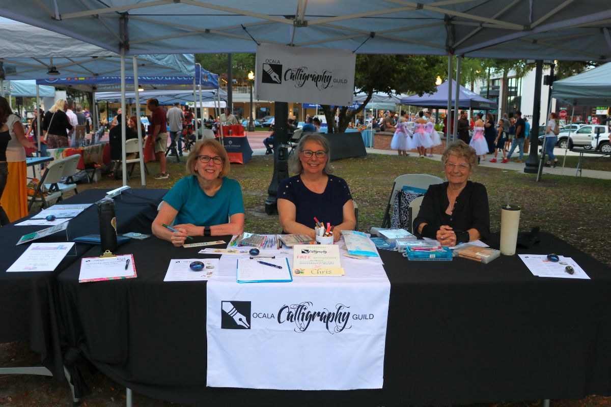 Three women sit behind a black table with a white table runner reading, "Ocala Calligraphy Guild." The table featured sign-up sheets and calligraphy activities.