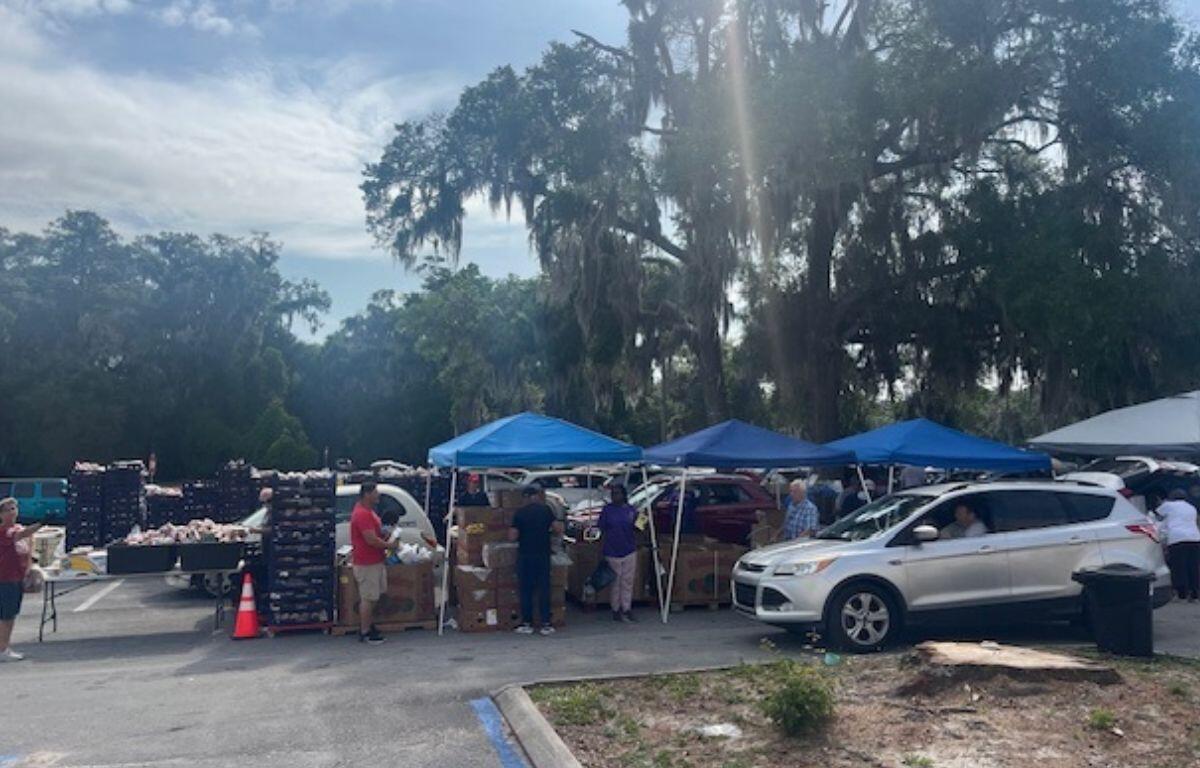 On the far left, palettes of food, trees in the background, people and cones in front of the palettes, and to their right, tents, and in front of that a car, with people handing out bags of food to people in cars.