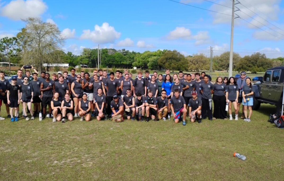 Background, trees, utility pole, middle a group of people to the extreme right a pickup truck, and in the foreground a grass field.