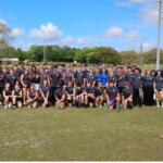 Background, trees, utility pole, middle a group of people to the extreme right a pickup truck, and in the foreground a grass field.