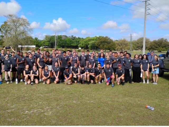 Background, trees, utility pole, middle a group of people to the extreme right a pickup truck, and in the foreground a grass field.