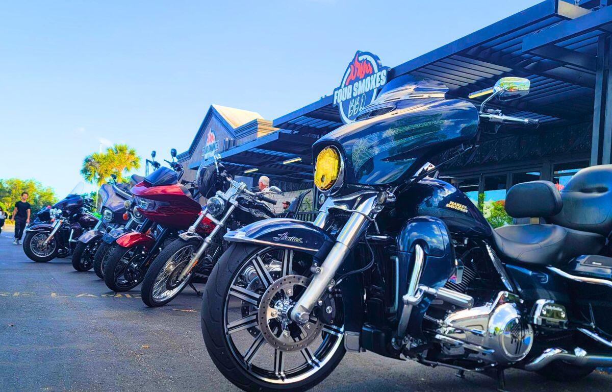 Motorcycles are lined up in front of a restaurant on a road. The restaurant features a sign above it reading, "Four Smokes BBQ."