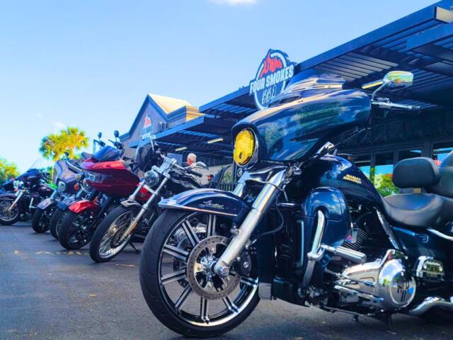 Motorcycles are lined up in front of a restaurant on a road. The restaurant features a sign above it reading, "Four Smokes BBQ."