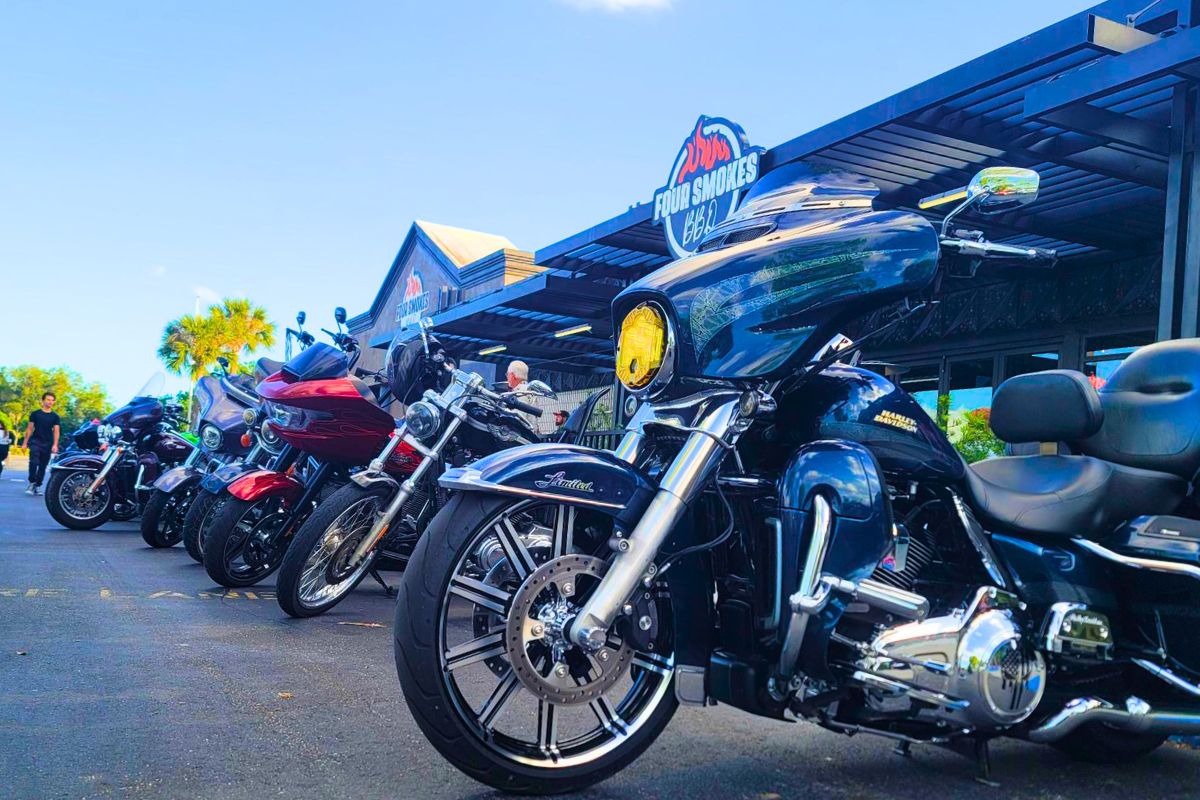 Motorcycles are lined up in front of a restaurant on a road. The restaurant features a sign above it reading, "Four Smokes BBQ."