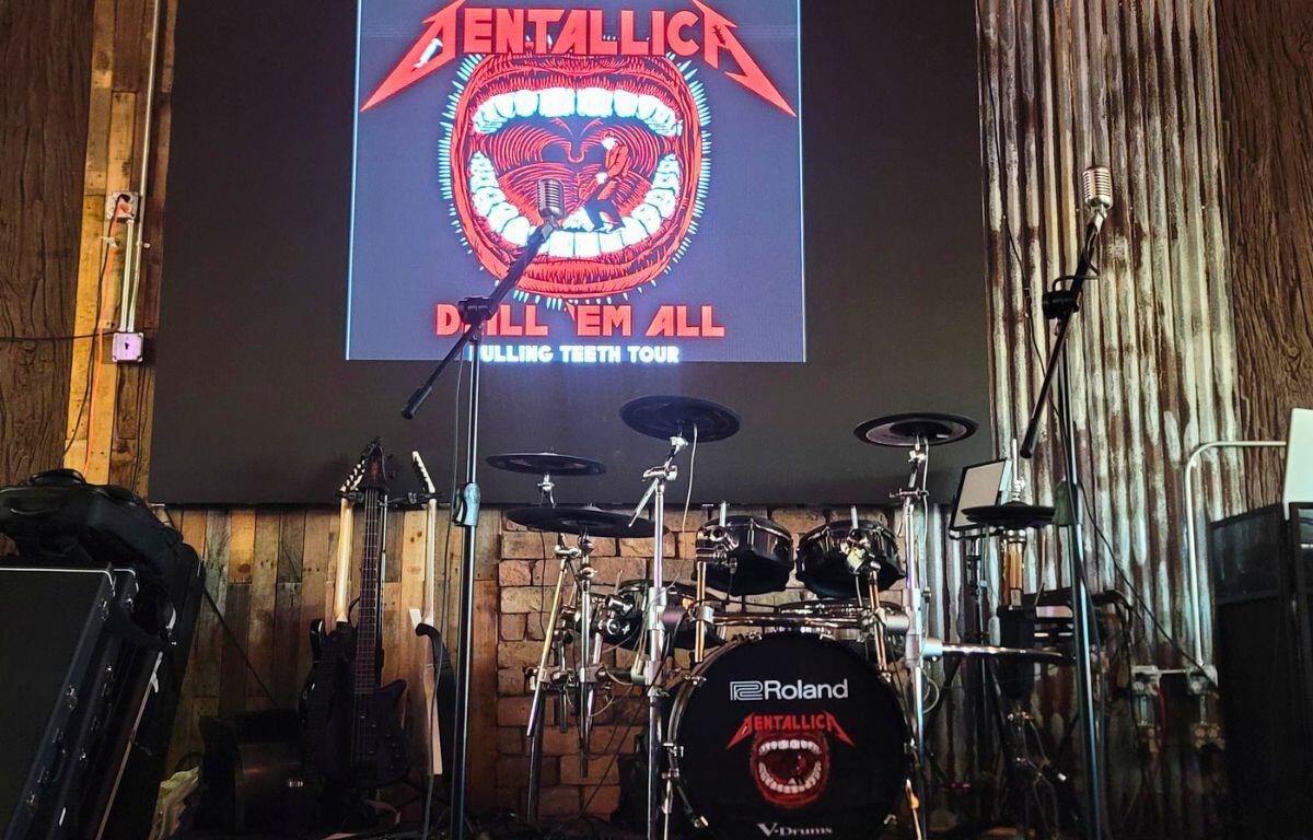 A drum set and two metal microphones are set on a platform with a large screen in the background. On the screen is a logo with a person drilling a mouth full of teeth and text around it that reads, "Dentallica. Drill 'Em All. Pulling Teeth Tour."
