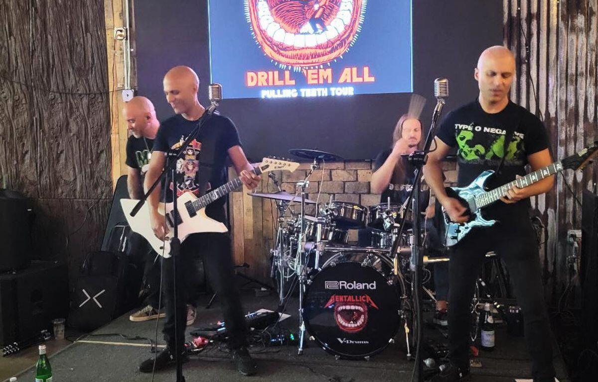 A band smiles and focuses while playing music on a platform. In the background is their logo on a large screen with a mouth full of teeth with a man drilling into it.