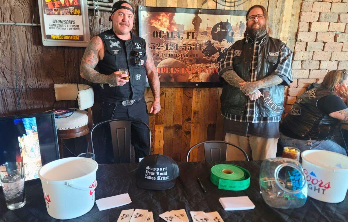 Two men stand in front of a poster advertising a tattoo shop in a restaurant setting. In front of them is a table with business cards and raffle tickets.