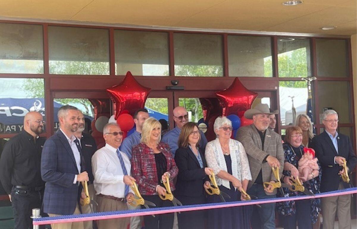 A sea of people standing in front of a multicolored ribbon. The people are holding large scissors. In the background is the front of a building.