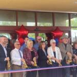 A sea of people standing in front of a multicolored ribbon. The people are holding large scissors. In the background is the front of a building.