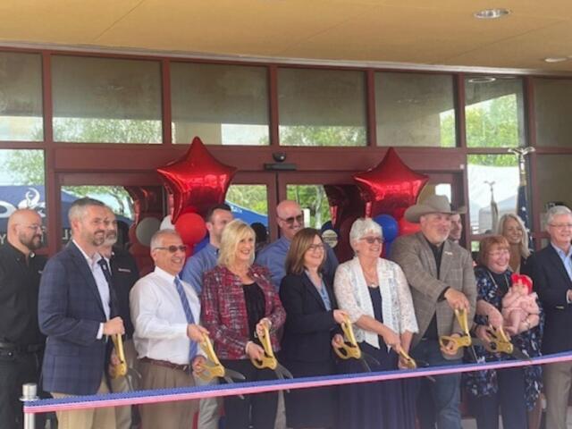 A sea of people standing in front of a multicolored ribbon. The people are holding large scissors. In the background is the front of a building.