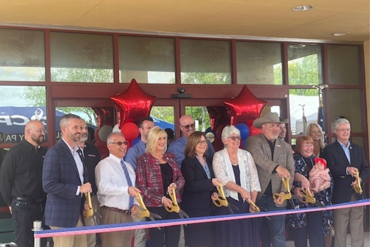 A sea of people standing in front of a multicolored ribbon. The people are holding large scissors. In the background is the front of a building.