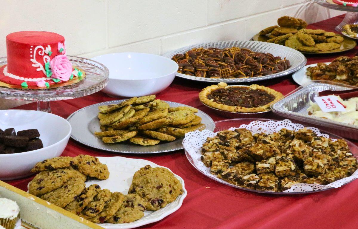 Dessert table with a pink frosted cake and trays of cookies, brownies, and bars on a red tablecloth.