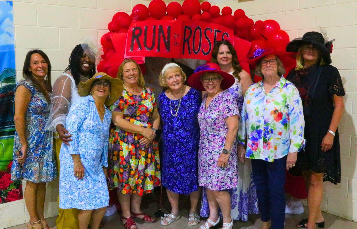 A group of women in colorful dresses posing under a red balloon arch that reads 'RUN for the ROSES'.