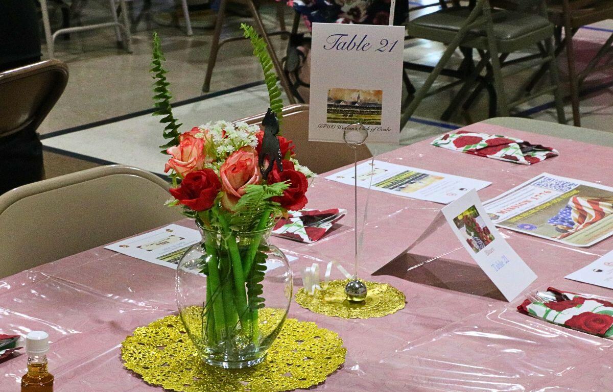 A clear, glass vase with flower pens inside of it sits on a circular gold matt on top of a pink tablecloth. Near it is a card on a similar, but smaller gold matt reading, "Table 21." Flyers and napkins are scattered on the table.