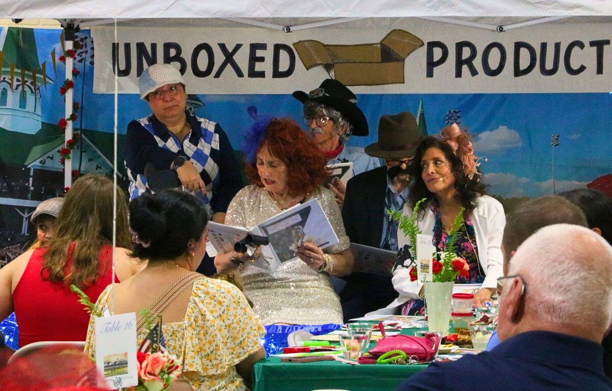 A crowd of guests watch costumed actors read lines from a script under a canopy. Behind the actors is a sign reading, "Unboxed Productions."