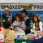 A crowd of guests watch costumed actors read lines from a script under a canopy. Behind the actors is a sign reading, "Unboxed Productions."