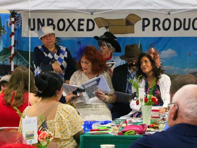 A crowd of guests watch costumed actors read lines from a script under a canopy. Behind the actors is a sign reading, "Unboxed Productions."