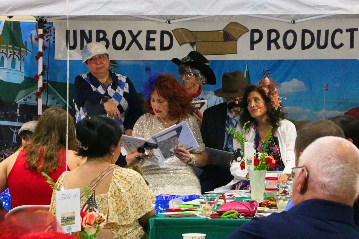 A crowd of guests watch costumed actors read lines from a script under a canopy. Behind the actors is a sign reading, "Unboxed Productions."