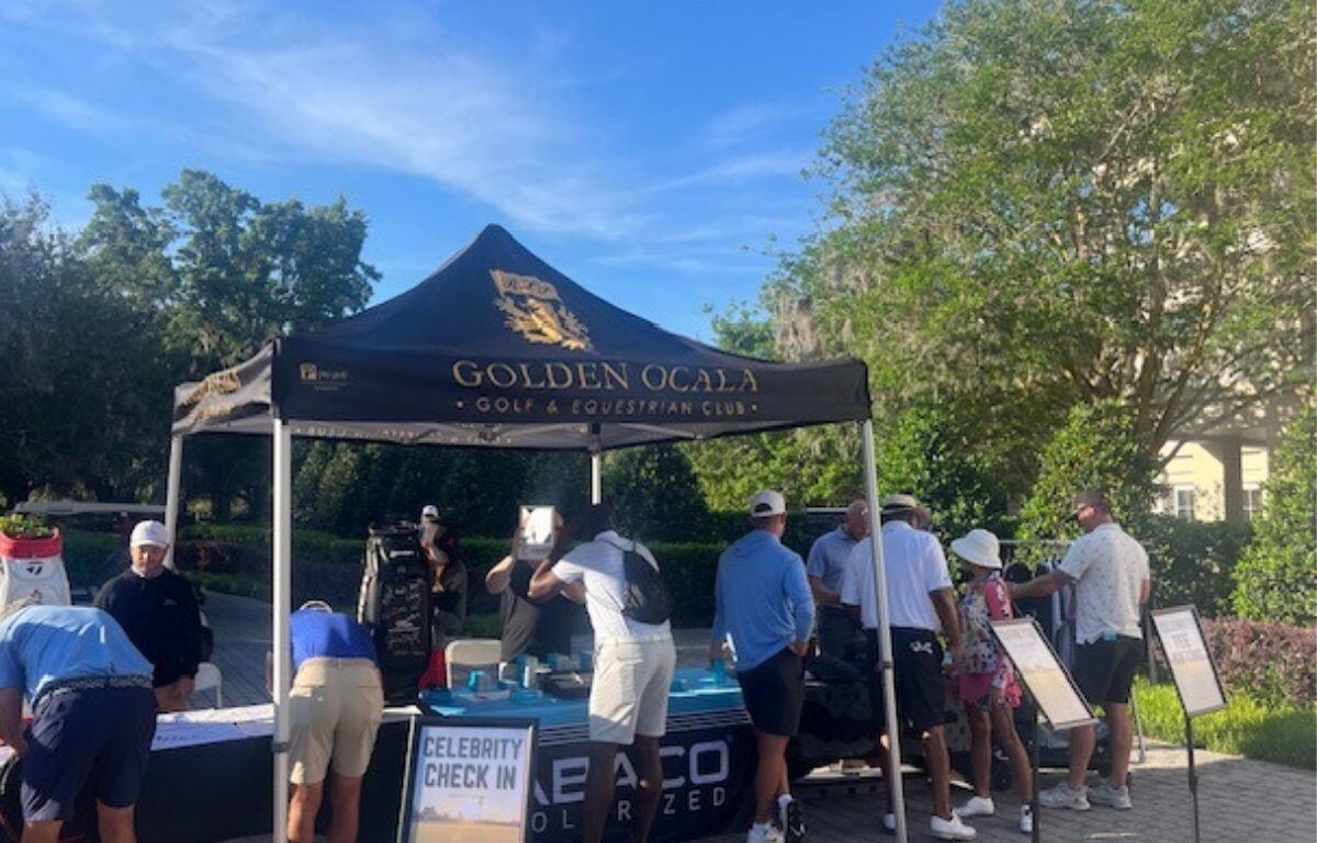 A tent with people standing behind a table, people in front signing papers, a placard with wording in front, tree line in the background.