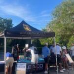 A tent with people standing behind a table, people in front signing papers, a placard with wording in front, tree line in the background.