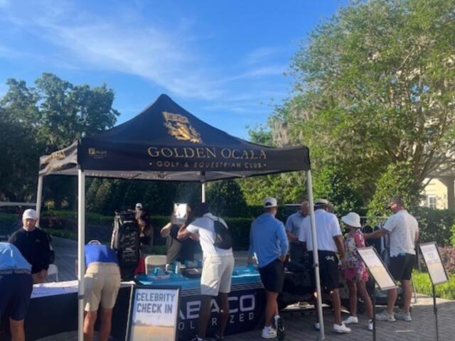 A tent with people standing behind a table, people in front signing papers, a placard with wording in front, tree line in the background.