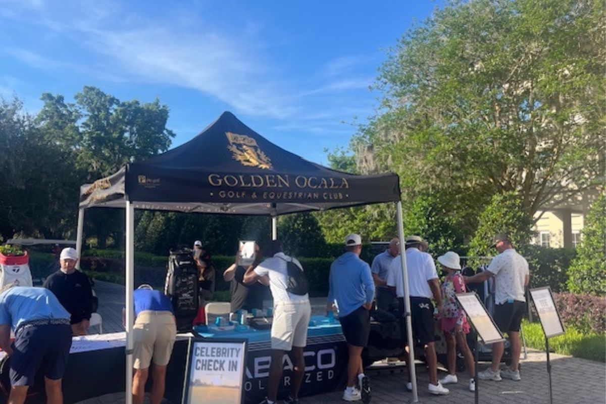 A tent with people standing behind a table, people in front signing papers, a placard with wording in front, tree line in the background.