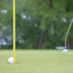 A golf ball is on the edge of falling into a hole with a flag. In the background are golf clubs and other golf balls.