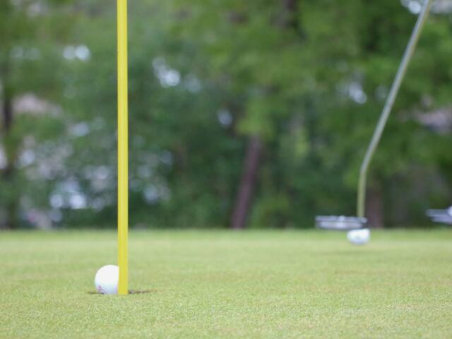 A golf ball is on the edge of falling into a hole with a flag. In the background are golf clubs and other golf balls.