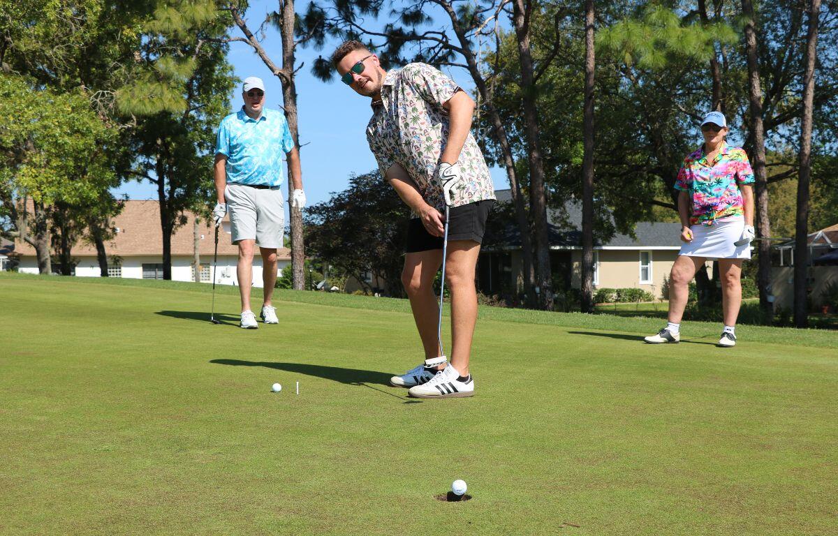 Golfer lines up a putt on a sunny green while two spectators stand nearby with trees in the background.