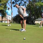 Golfer lines up a putt on a sunny green while two spectators stand nearby with trees in the background.