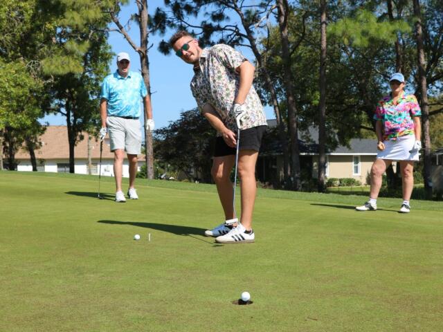 Golfer lines up a putt on a sunny green while two spectators stand nearby with trees in the background.