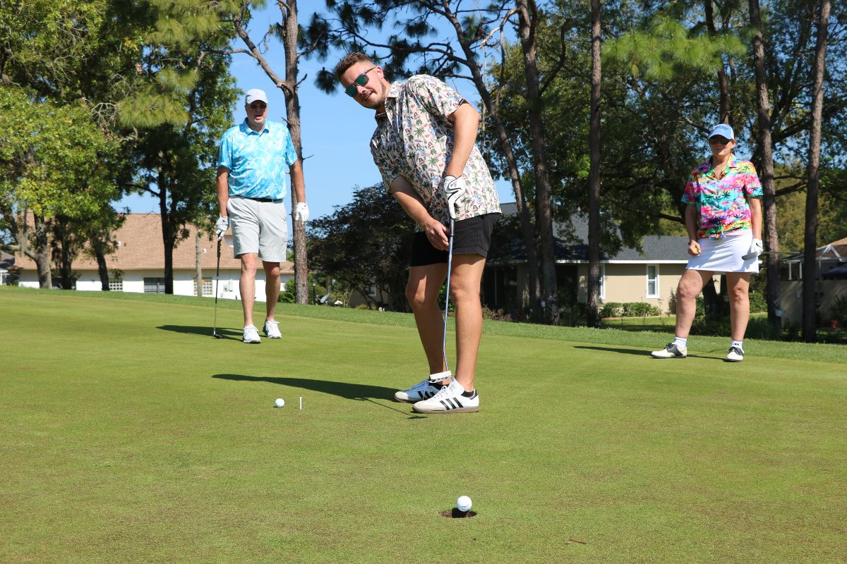 Golfer lines up a putt on a sunny green while two spectators stand nearby with trees in the background.