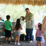A man demonstrates a tortoise and snake to children and families under a chickee.
